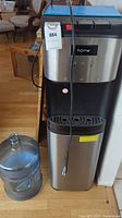 Full view of stainless steel HomeLabs water dispenser unit with attached power cord and 5-gallon water jug placed beside it on the floor.