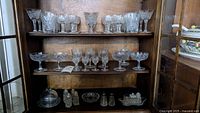 Wooden shelving unit displaying crystal glassware including wine glasses, champagne coupes, tumblers, salt and pepper shakers, and small glass service ware items
