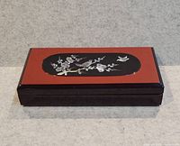 Closed rectangular lacquer jewelry box on neutral background showing front and top with inlaid bird and floral design.
