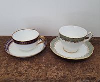 Two decorative teacups with matching saucers on wooden surface, one with blue and gold detailing and the other with teal and gold pattern.