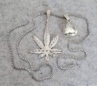 Two silver pendants laid flat on grey felt background with thin box chain necklaces. One pendant is a detailed cannabis leaf, the other a hammered heart shape.