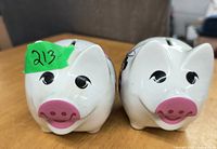 Front view of two ceramic piggy banks side by side, showing pig snouts, eyes, and coin slots on top.