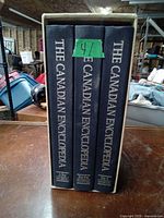 Three hardcover books standing upright with titles 'The Canadian Encyclopedia' visible on spines. Middle volume is smaller and has a green numbered label.