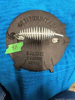 Top view of the cast iron bacon press showing coil spring handle and raised lettering, placed on blue cloth.