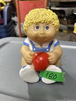 Photo of Cabbage Patch doll coin bank showing front view with yellow curly hair, blue eyes, blue and white outfit, and red ball held in hands.
