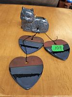 Photo of a ceramic cat figurine resting on a table with three heart-shaped ceramic decor pieces glazed in brown and black with string loops for hanging