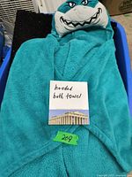 Photo of a teal child's hooded bath towel with shark face on the hood, laid out on a surface with a handwritten note 'hooded bath towel' and green lot number '209'.