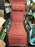 Gravity Lounge Chair with red fabric seat and black armrests, padded headrest pillow attached, shown upright surrounded by miscellaneous storage items.