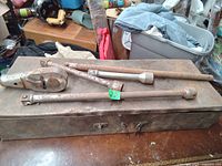 Metal ratchet wrench and three socket extension bars placed on a closed metal box, showing surface rust and signs of wear.