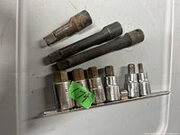 Top-down image of seven hex tools on a white surface including two rusty hex socket extension bars, two black hex socket extensions, and three chrome hex sockets.