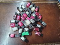 Lot of 29 small sealed nail polish bottles in assorted colors including metallic silver, pink, red, white, and plum on a brown textured surface.
