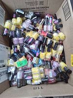 Photo showing a cardboard box filled with numerous small nail polish bottles in mixed colors including yellow, pink, purple, and red. Each bottle has a black cap, all appear sealed and unused.
