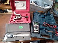 Photo showing four tools: an OEM valve spring compressor in red case, Motorcraft digital caliper in black case, digital multimeter with leads in blue case, and blue-handled pliers.