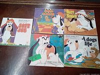 Photo of five vintage Droopy children's books arranged on a dark wood surface. Covers feature colorful cartoon illustrations of Droopy the dog in various themes. Titles include Soft Job, Dog Show, Bare Bones, Wise Old Dog, and A Dog's Life.