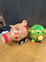 Photo showing the pink piggy bank on wheels with red snout and flower decoration, beside the green piggy bank with orange hooves.