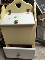Front view of yellow wooden potato storage bin with white lifting flap door and bottom drawer open, showing wooden knobs and heart cutout design.