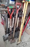 Front view of square end shovels with wooden shafts and red D-grip handles, alongside magnetic rakes and additional shovels.