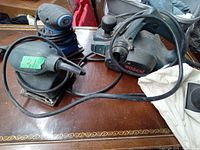 Three power tools on a wooden table: two sanders and one Bosch electric planer. Cords are attached, tools show used condition.