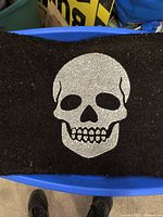 Top view of rectangular black doormat with silver skull graphic in center, showing texture and design details.