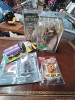 Photo of four collectible toy items on a table: McFarlane Army Shock Trooper figure, male figure in suit, purple toy car, and fingerboard skateboard with clown figure in packaging.