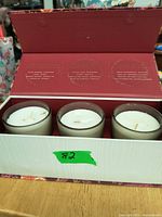 Three Vanilla Bourbon scented candles in glass containers inside open rectangular box with red interior and light exterior. Candles are unused and the box has scent descriptions.