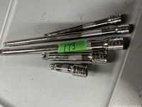 Six metal socket extension bars with knurled grips on a gray tray, varying lengths, marked with green tape '173'