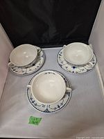 Three white porcelain soup cups with two handles and matching saucers, decorated with blue floral and vine patterns, set against a neutral background.