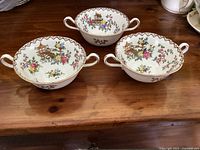 Three cream soup bowls with double handles, decorated with Pagoda and floral pattern, placed on wooden surface.