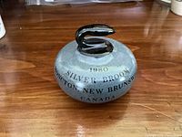 Front view of the souvenir miniature curling stone showing text 1980 Silver Broom Moncton New Brunswick Canada with a black handle on top.