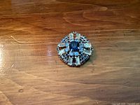 Front view of a round brooch pin with a central blue stone, surrounded by clear rectangular and small blue round stones set in silver-tone metal on a wooden surface.