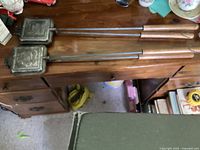 Two vintage Coghlan's sandwich camp cookers with long handles and wooden grips, shown on a wooden surface.