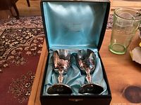 Two silver plated goblets engraved 'Mom' and 'Dad' in original box with blue satin lining, shown on wooden surface with carpet background.