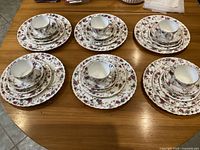 Photo showing the full set arranged in six place settings, each consisting of 5 pieces: cup, saucer, small plate, medium plate, and large plate all featuring floral design with gold edging.