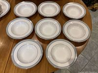 Eight rimmed soup bowls arranged on a wooden table showing white base color with a light blue band and silver rim.