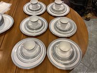 Six place settings arranged on a wooden table, each setting includes five pieces: cup, saucer, bread plate, salad plate, and dinner plate with intricate silver and grey patterns along rim