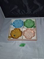 Four colored cloverleaf-shaped glass dishes in a box separated by white dividers. Colors are light blue, green, amber, and pink. Each dish has a ruffled edge and a quilted pressed pattern.