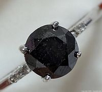 Close-up photo showing the round black diamond center stone set on a 14KT white gold band with small white diamond accents.