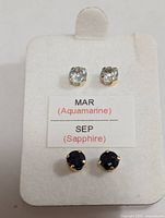 Front view of white display card showing both pairs of stud earrings with one pair light blue aquamarine stones and one pair dark blue sapphire stones along with MAR (Aquamarine) and SEP (Sapphire) labeling.