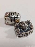 Two silver beads displayed on a white background. One is cube-shaped with letters and a gold heart accent, the other is a detailed domed building shape.