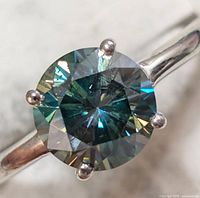 Close-up of round blue moissanite gemstone set in silver ring band, showing color and facets.