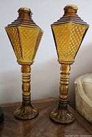 Pair of tall amber glass candle holders with lantern designs and detailed bases, shown side by side.