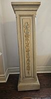 Full view of tall rectangular antique white pillar with gold painted decorative vertical floral design, showing entire piece with base and top shelf.