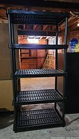 Black plastic 5-shelf storage unit standing upright in unfinished room showing full shelving structure.
