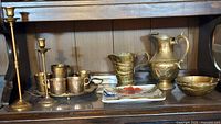 Full view of the brass items with candlesticks, cups, jugs, hammered bowl and ceramic plate on wooden shelf