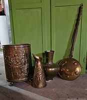 Four brass decorative items including waste bin, tall vase, pitcher, and bed warmer with wooden handle.