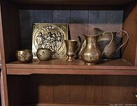 Brass decor items on a wooden shelf including candle holders, plaque, goblet cup, and pitchers.