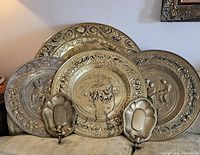 Four round brass decorative plates with ornate embossed designs arranged leaning on a surface with two brass wall sconces candle holders placed at front.