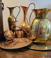 Copper and brass jugs, cup, piggy bank and decorative plate arranged on wooden surface.