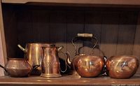 Front view showing six copper items including teapots, pitchers, and pots on a wood shelf