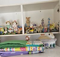 Cabinet shelf with multiple bunny figurines and bunny casserole dish alongside folded colorful Easter and spring-themed tablecloths.